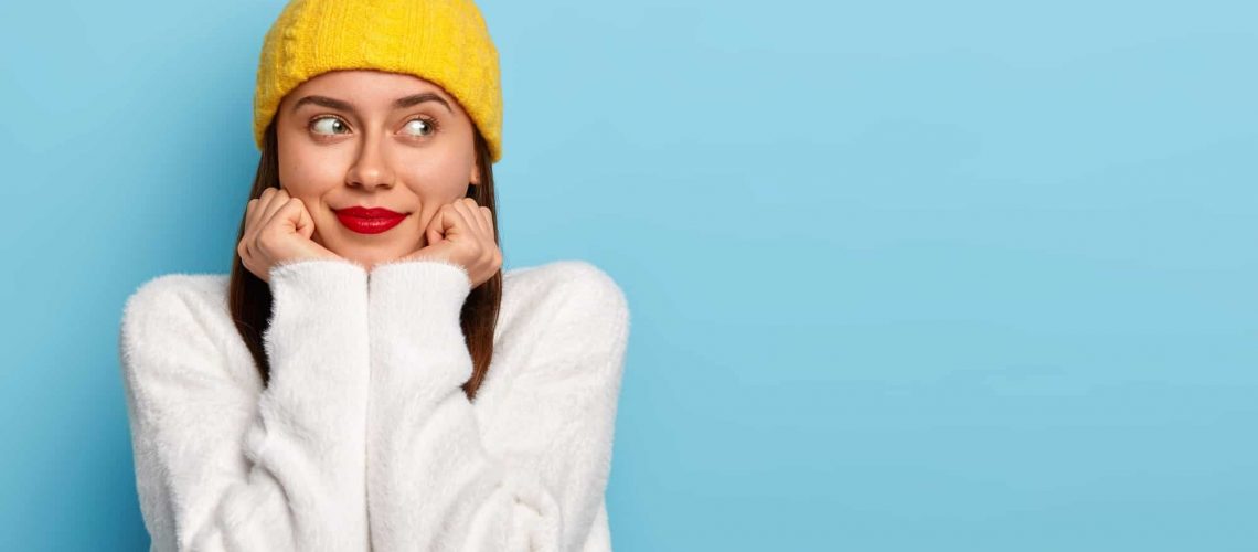 horizontal-shot-happy-dreamy-european-woman-wears-minimal-makeup-red-lipstick-looks-aside-dressed-yellow-hat-white-sweater-poses-against-blue-background-being-fascinated-pleased